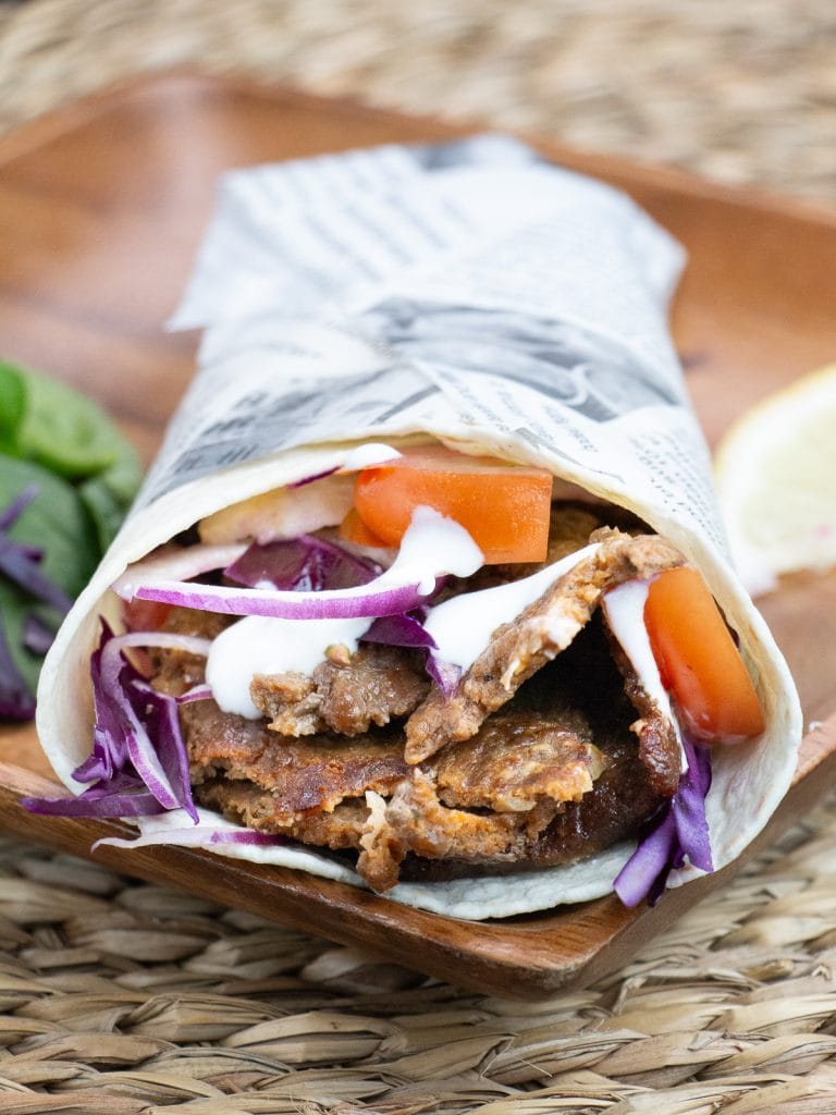Homemade doner kebab wrap with spiced beef, red cabbage, tomato, red onion and garlic yogurt sauce, wrapped in newspaper-style paper and served on a wooden board
