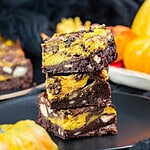 Stack of three pumpkin cheesecake brownies with chopped cashews and an orange swirl, on a black plate beside mini pumpkins