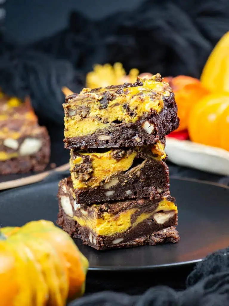 Stack of three pumpkin cheesecake brownies with chopped cashews and an orange swirl, on a black plate beside mini pumpkins