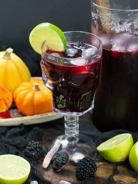Chicha morada witch’s brew in a skull glass with lime, blueberry eyeball ice cubes and blackberries, next to mini pumpkins